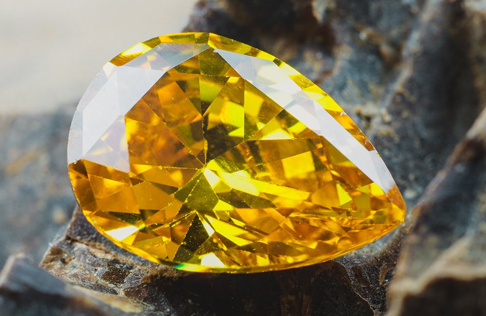 A yellow, pear-shaped gemstone lies on a textured, rocky surface, reflecting light with its multiple facets.