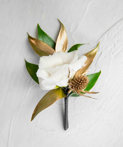 This boutonniere features an elegant ivory bloom paired with greenery in accents in gold